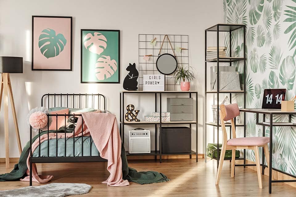Framed posters on a white wall above a cozy twin bed with pillows and blankets in a hipster teenager bedroom interior with leaves wallpaper