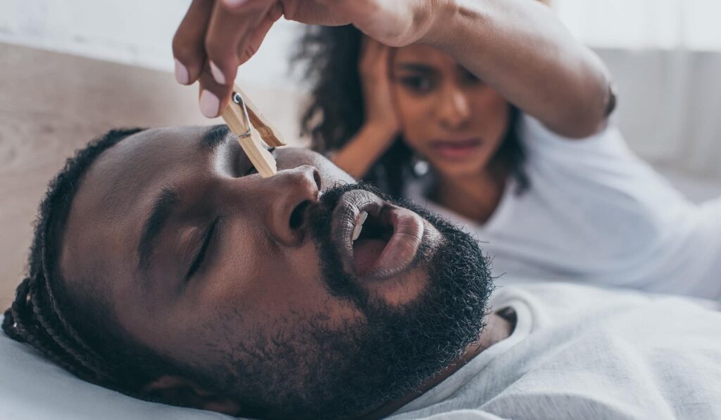 A woman in bed is inches away from closing a clothespin over her husband snores in his sleep next to her