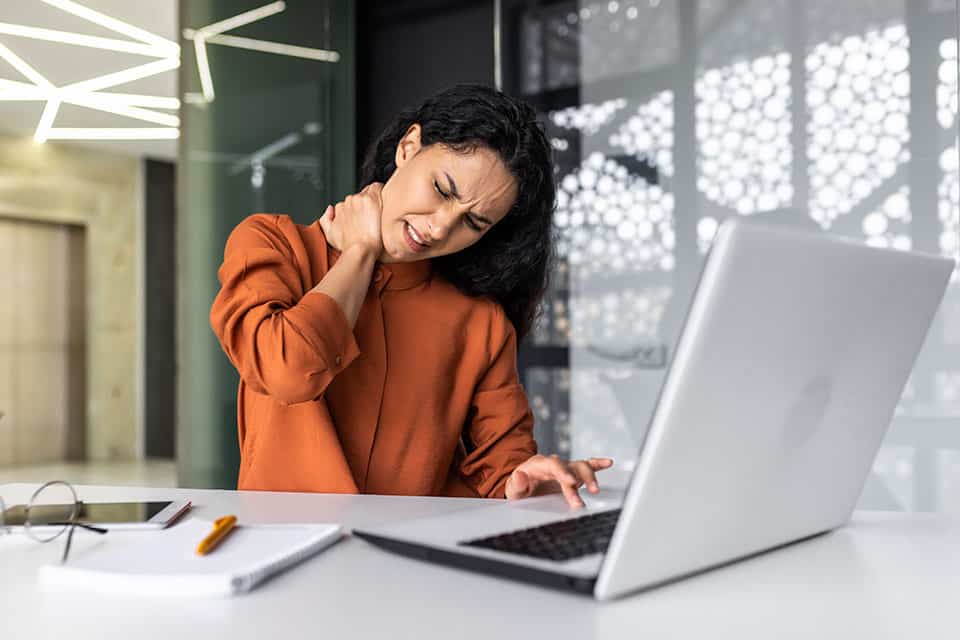 Overtired business woman working inside office with laptop and desk with bad ergonomics, Hispanic female worker has severe neck pain, massaging muscles with hand